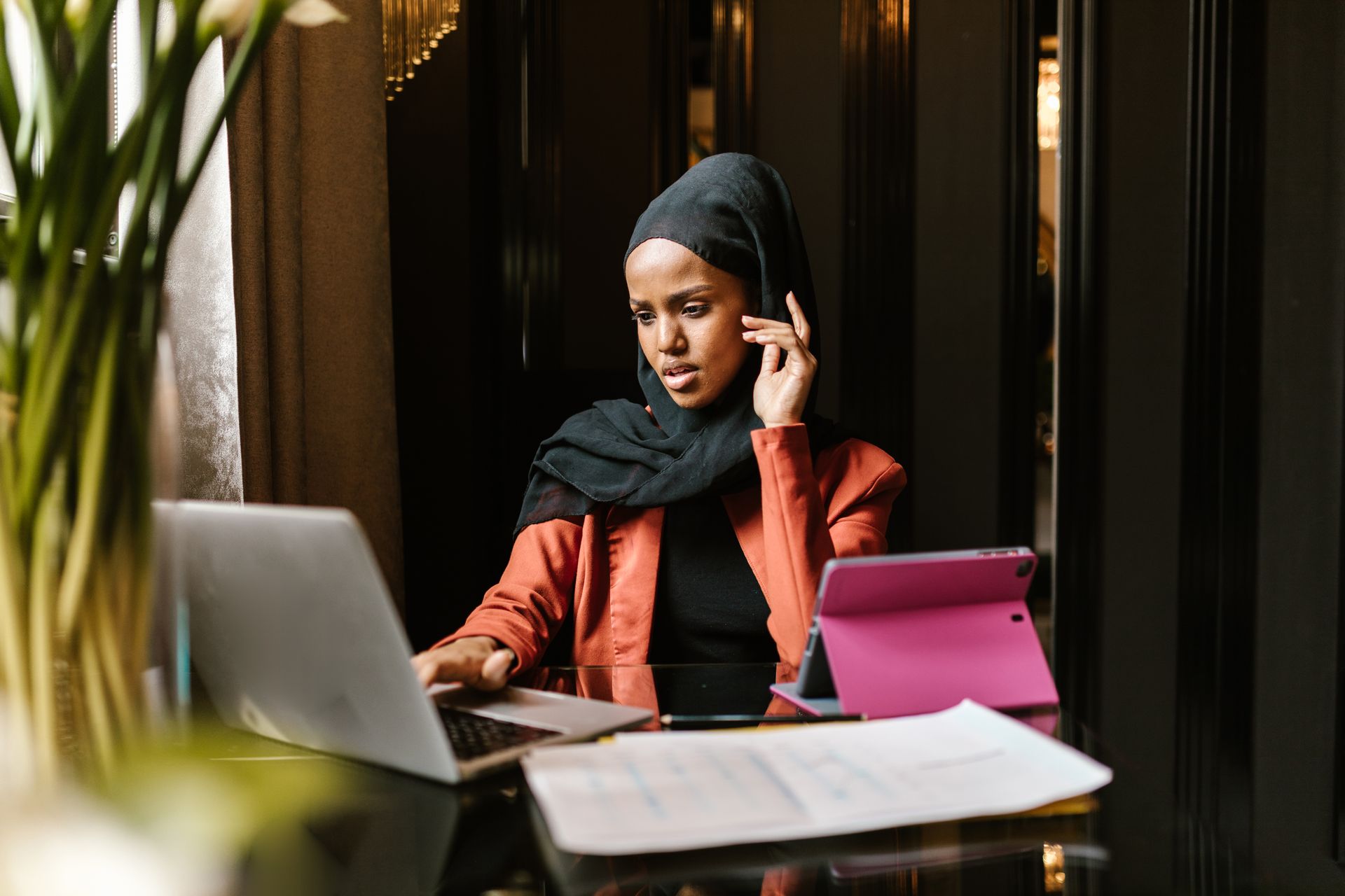 A Black businesswoman in a hijab reviewing figures on a laptop and tablet at her desk