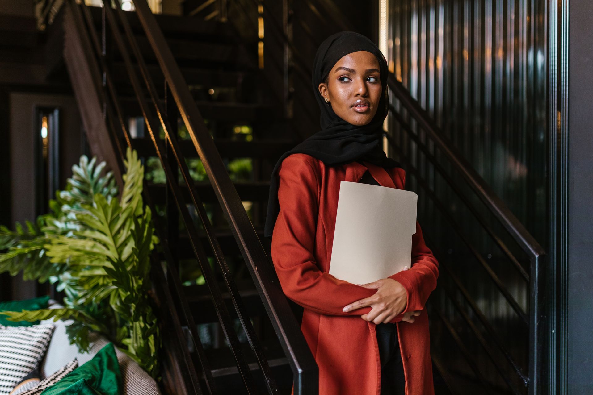 A Black businesswoman in a hijab and red suit carrying documents in an office